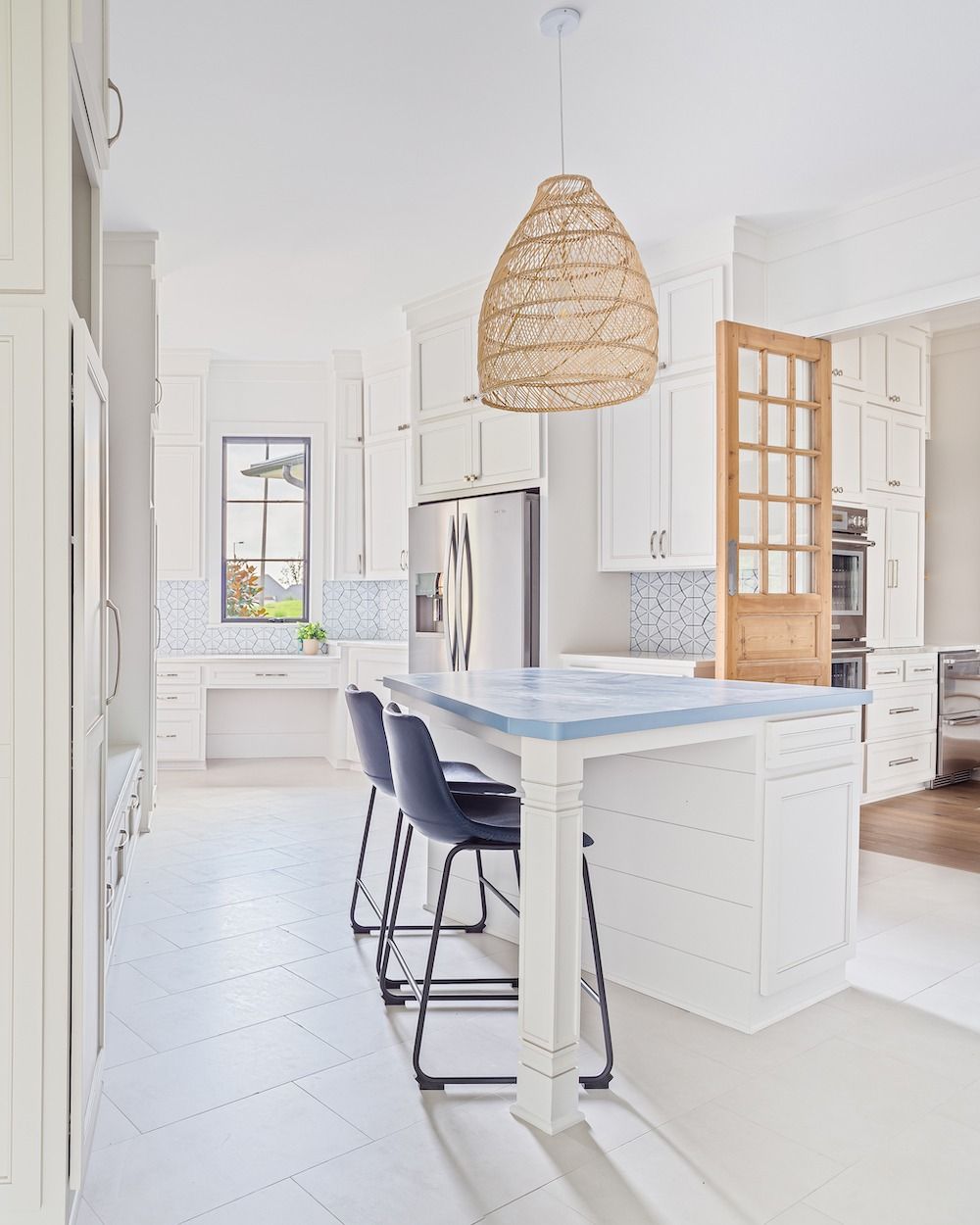 A bright, white kitchen featuring a central island with blue countertops, two navy bar stools, and a textured pendant light.