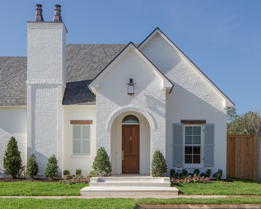 White brick house with a dark wood arched front door, grey shutters, and a chimney under a clear blue sky.