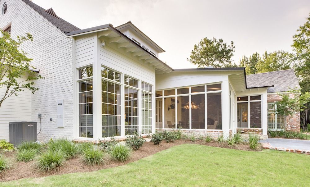 A white modern farmhouse with large windows and a screened-in porch, surrounded by green landscaping and trees.