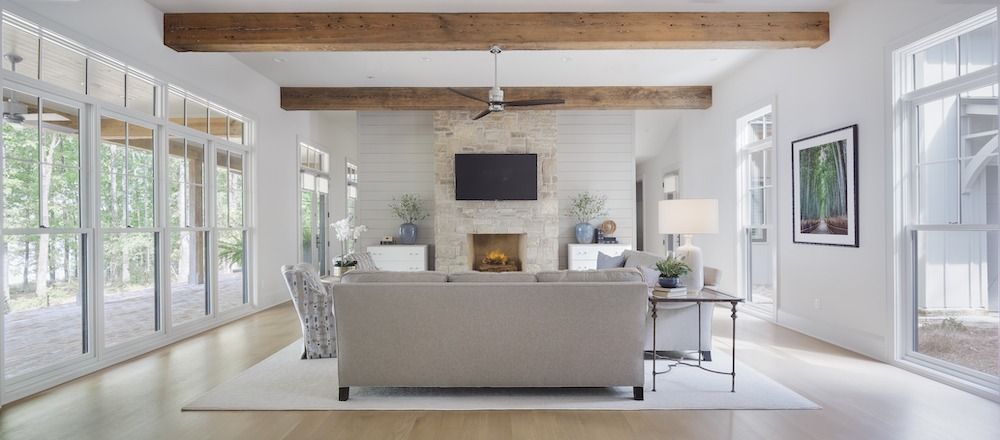 A bright living room with white walls, wooden ceiling beams, a stone fireplace, and a light-colored sofa facing a TV.
