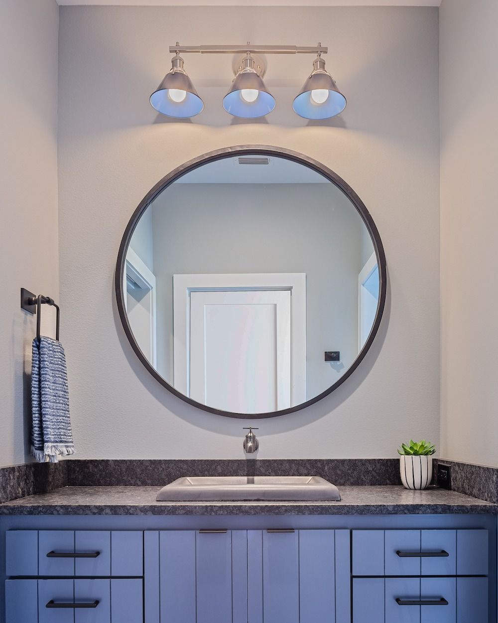 A round mirror hangs above a blue bathroom vanity with a countertop sink, a three-light fixture, and a towel on the left.