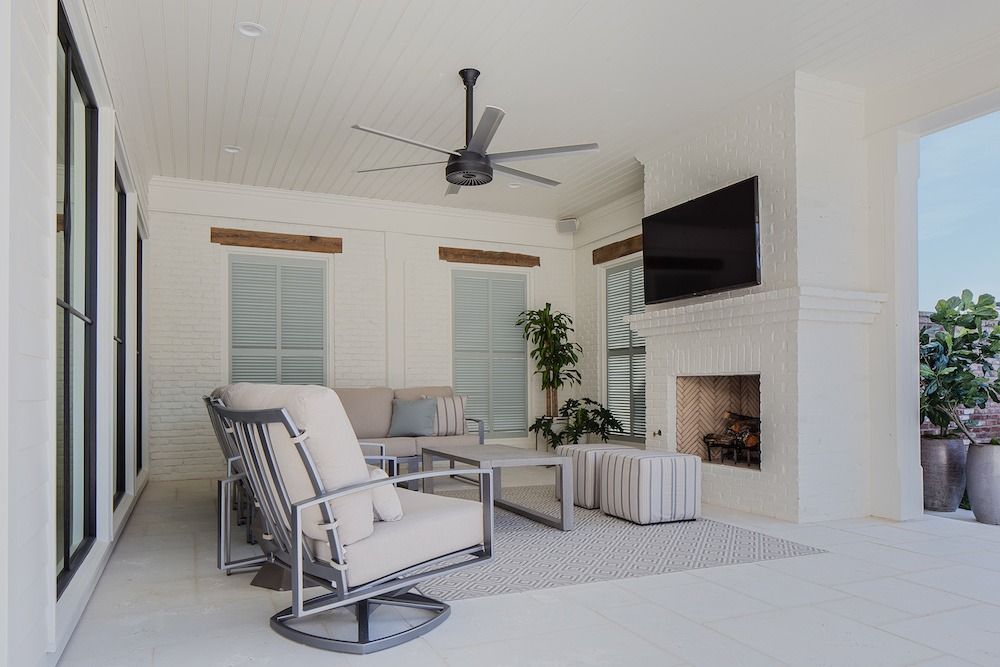 A covered patio with a white fireplace, wall-mounted television, comfortable furniture, and a ceiling fan.