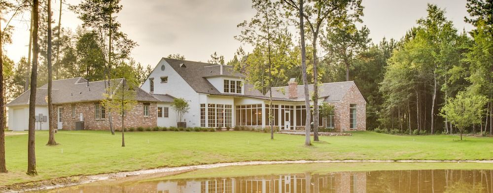 A multi-story house with white siding and brick accents sits on a grassy lawn beside a pond, surrounded by many trees.