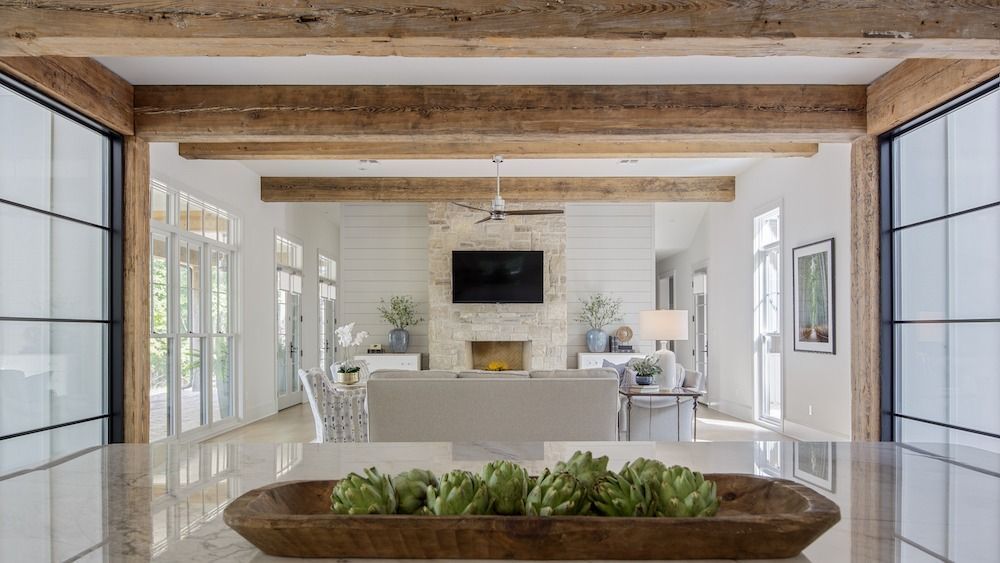A bright, airy living room with exposed wooden ceiling beams, a stone fireplace, and a bowl of artichokes in the foreground.