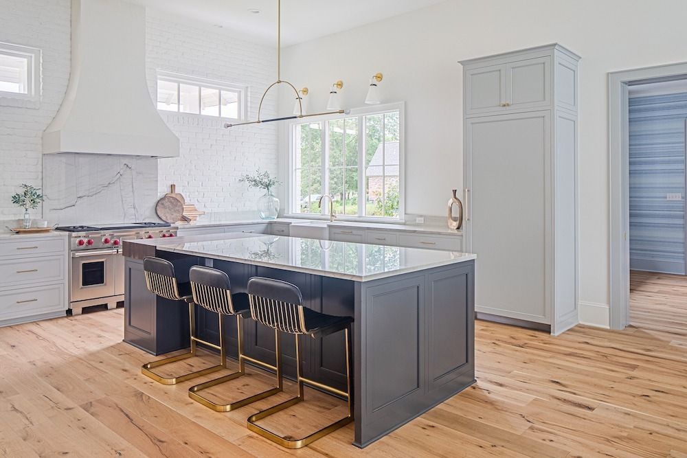 A bright kitchen with a large, dark-gray island, white marble counters, gold barstools, and light-colored hardwood floors.