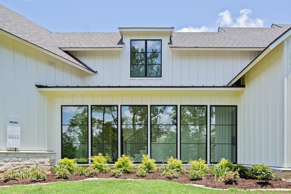 A modern white farmhouse with vertical siding, black-framed windows, a gray roof, and a low stone base in a grassy yard.