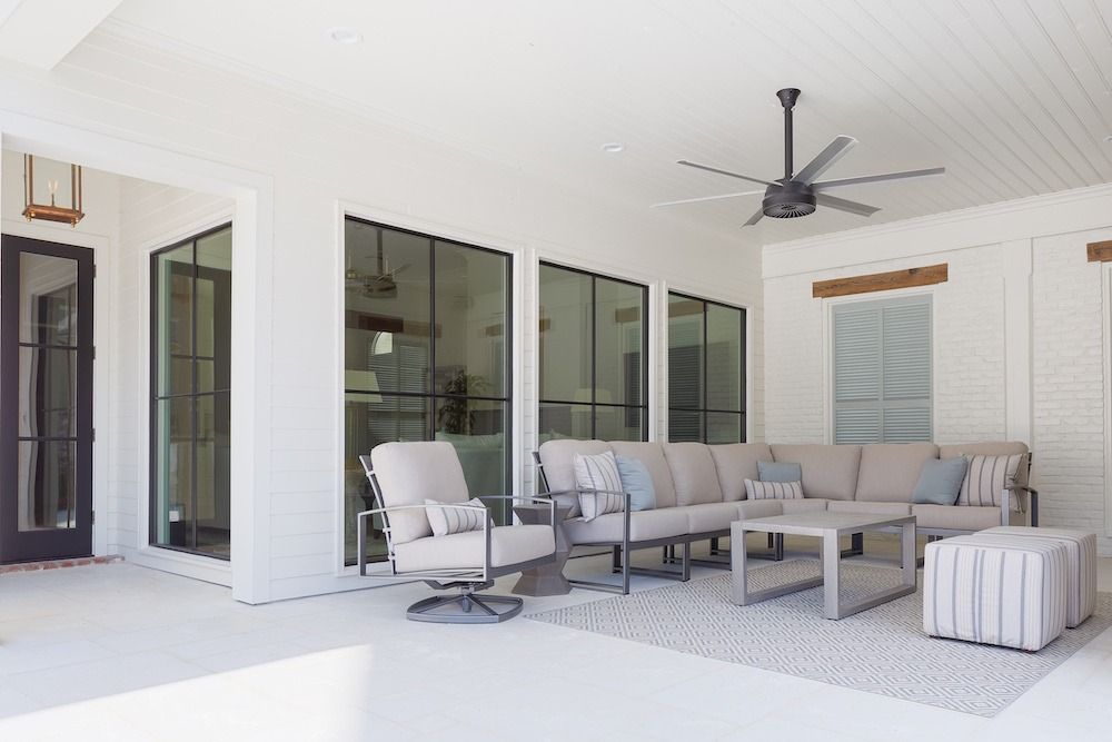 A bright, covered outdoor patio featuring a neutral sectional sofa, a swivel chair, two ottomans, and a ceiling fan.