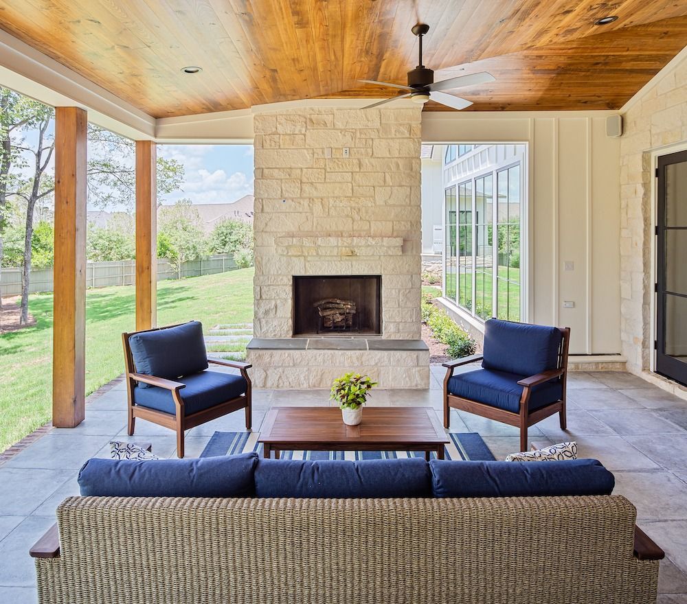 A covered patio features a stone fireplace, a sofa with blue cushions, two armchairs, and a wooden coffee table.