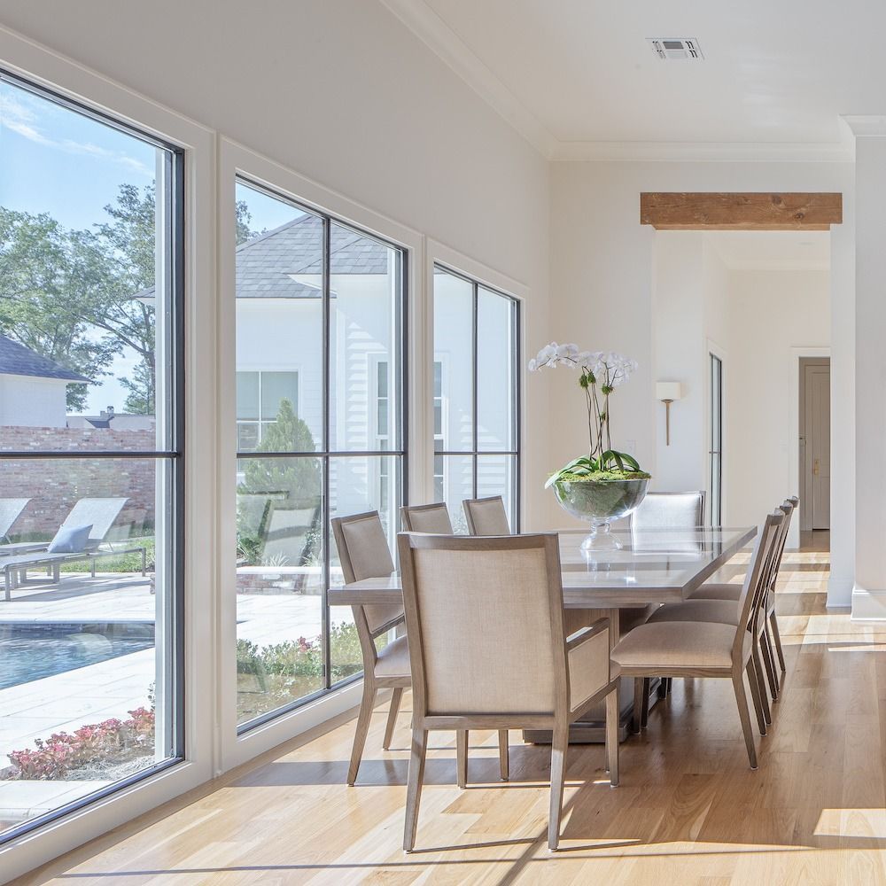 A modern dining room with light wood floors, a long table, and chairs overlooking a backyard pool through large windows.