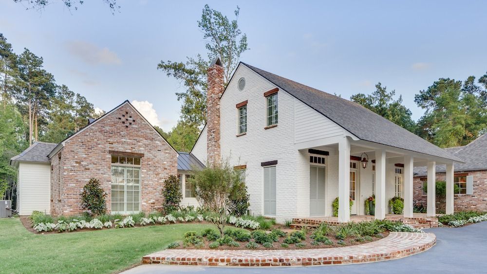 A rustic, two-story house with a brick-and-white facade, a columned front porch, and a manicured garden.