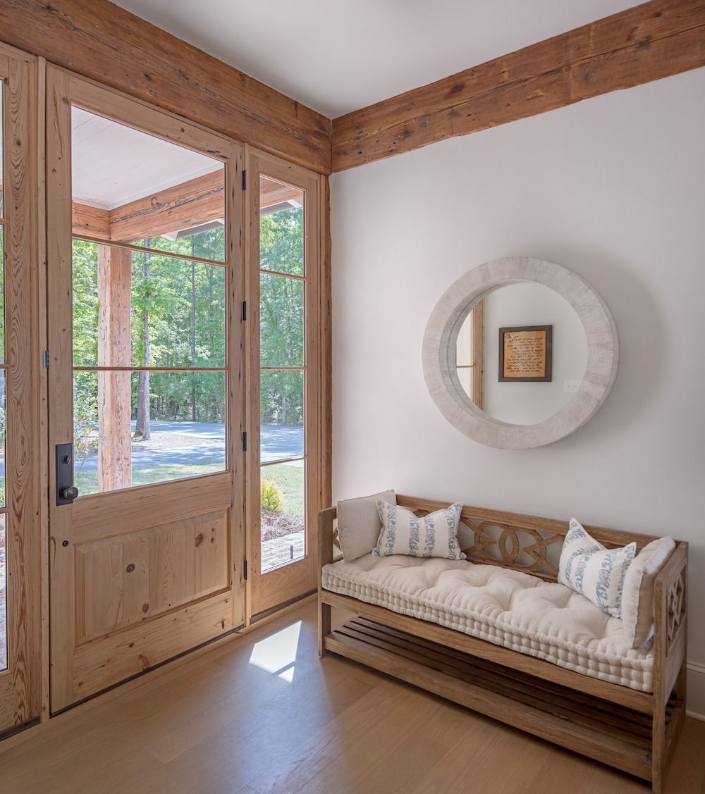 A wooden entryway with a bench, a round mirror on a white wall, and a large wooden door looking out to trees.