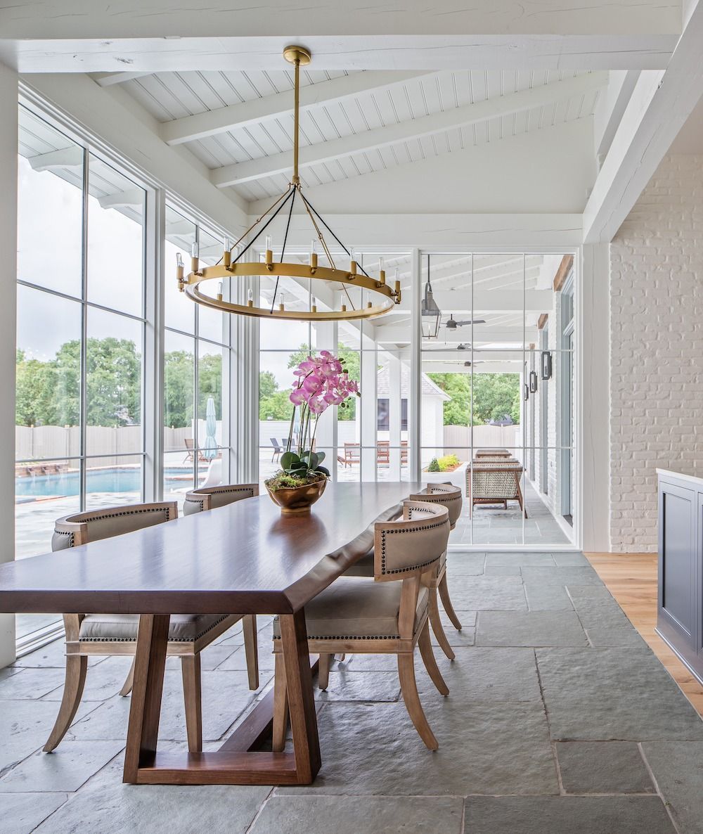 A dining area on a covered patio with a wooden table, upholstered chairs, and a large metal chandelier overlooking a pool.