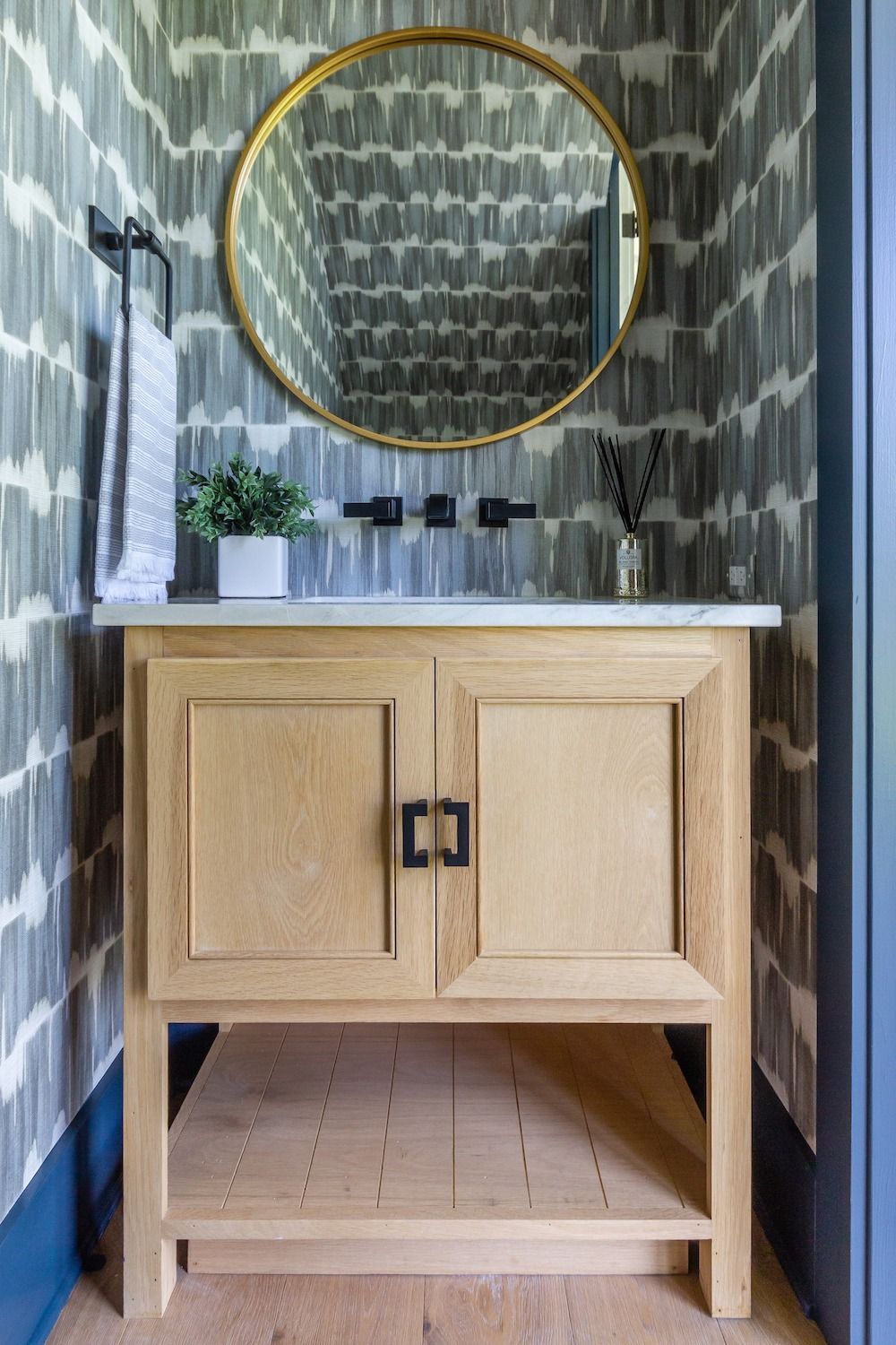 A wooden bathroom vanity with a white countertop and gold circular mirror against gray patterned wallpaper.