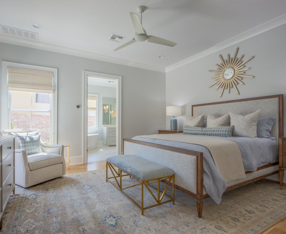 A bright bedroom with a king bed, upholstered headboard, a gold sunburst mirror, a ceiling fan, and a cozy armchair.