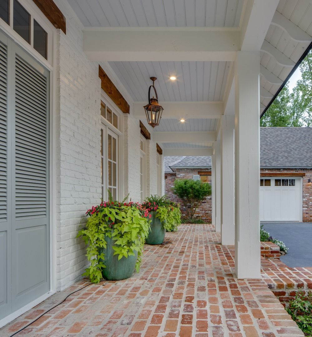A covered porch with a red brick floor, white brick walls, large green potted plants, and a hanging lantern light fixture.