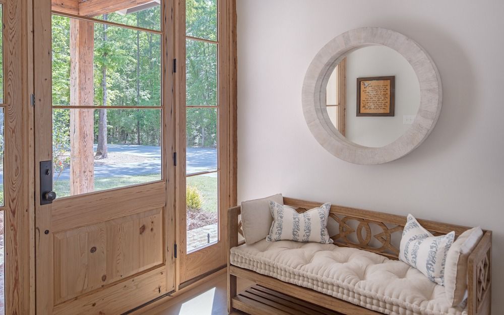 A wooden bench with cream cushions and pillows sits in a bright entryway next to a glass-paned door and round wall mirror.