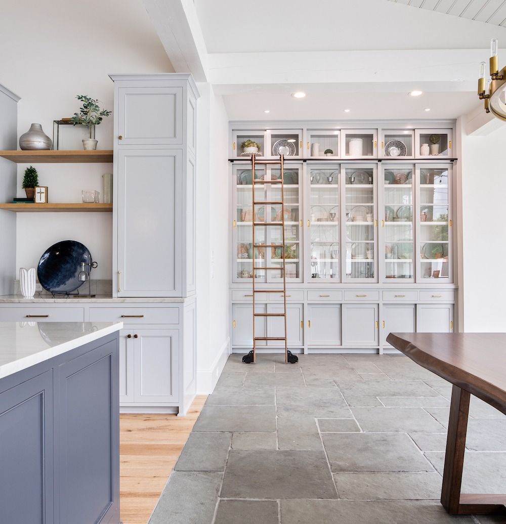 A bright kitchen features gray cabinetry, built-in display shelving, a wooden ladder, and a stone floor beside a table.