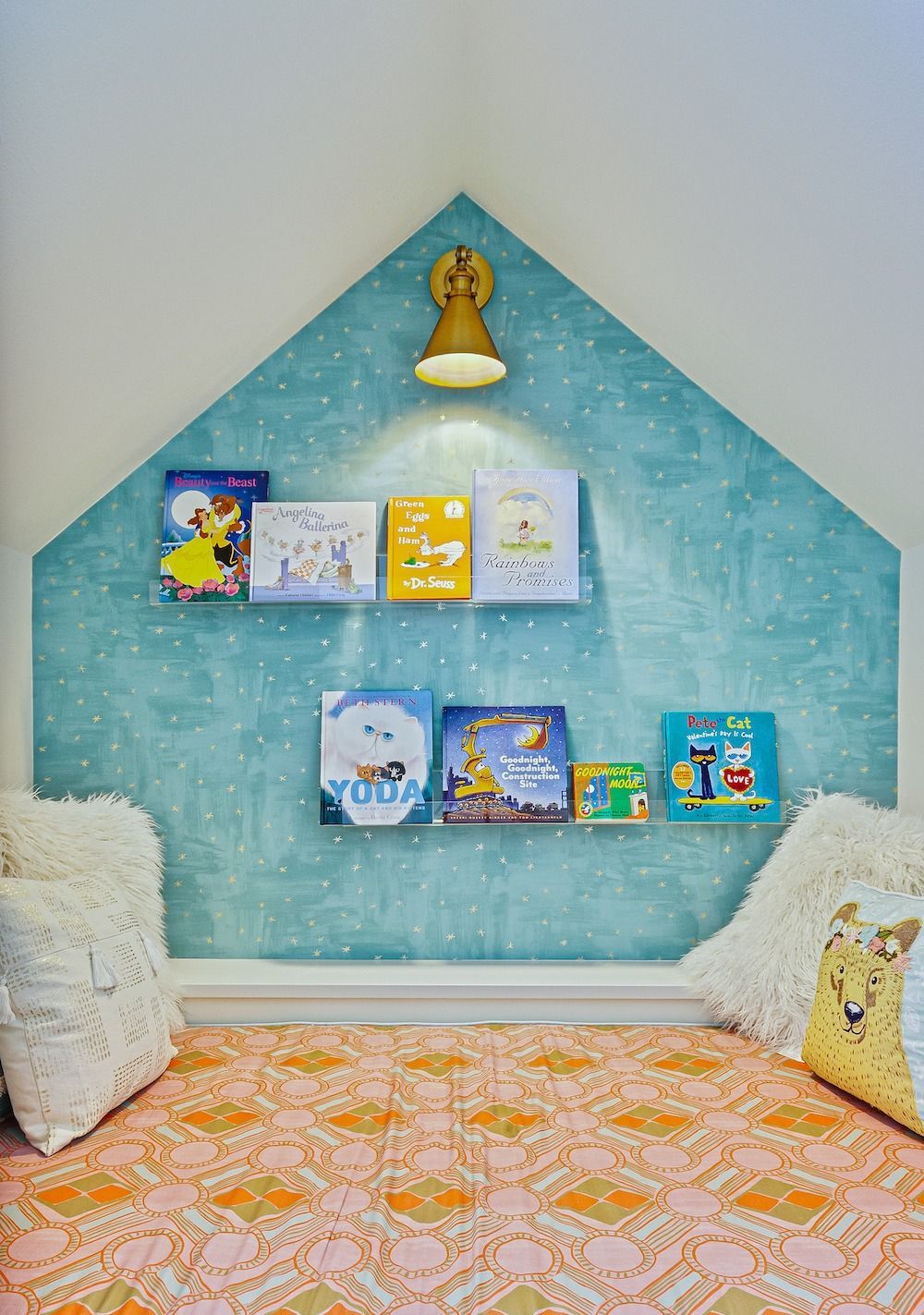 A cozy reading nook with a gabled blue star-patterned wall, two floating bookshelves, a gold lamp, and plush pillows.