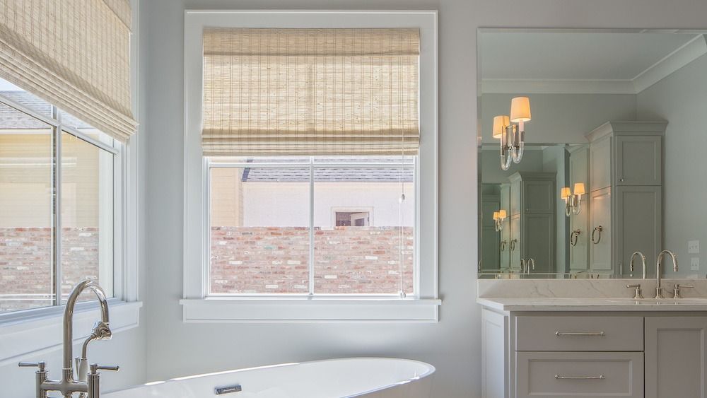 A bright bathroom featuring a white bathtub, a window with a bamboo shade, and a vanity with a mirror and light fixtures.