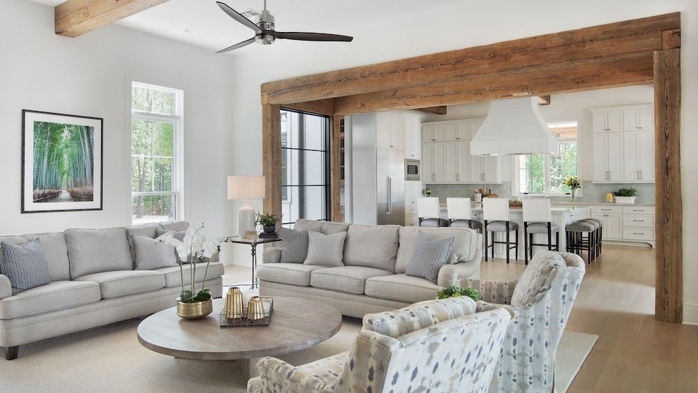 A bright, open-plan living room with light gray couches, a round coffee table, and an exposed wooden beam frame.