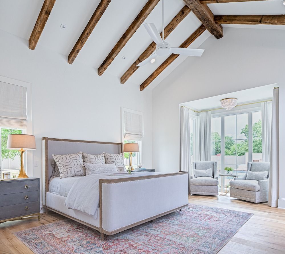 A bright bedroom features a light grey bed, vaulted ceiling with wood beams, a white ceiling fan, and two armchairs by a window.