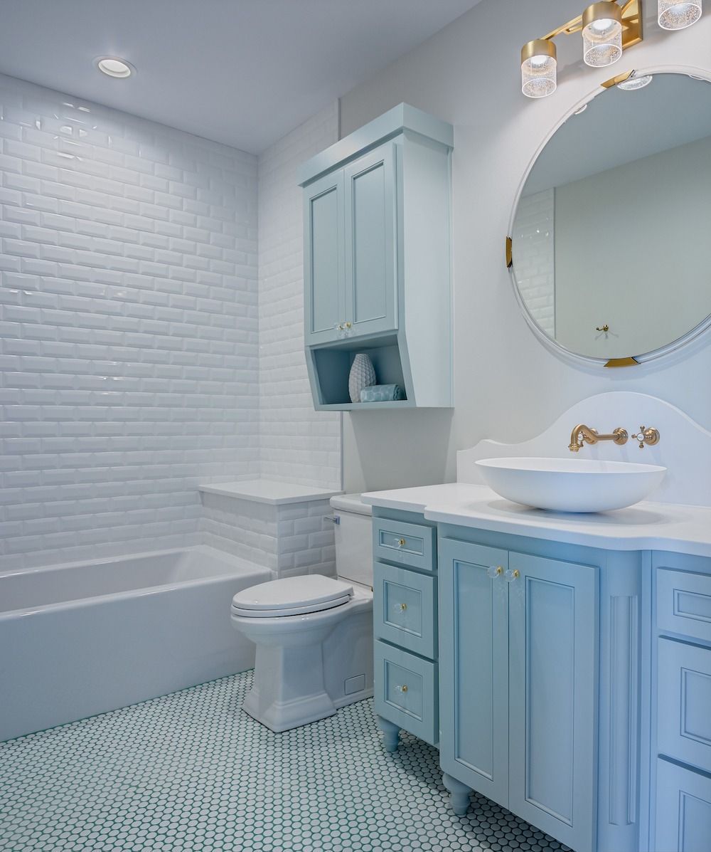 A modern bathroom with light blue cabinets, a white vessel sink, a circular mirror, and patterned blue and white floor tile.