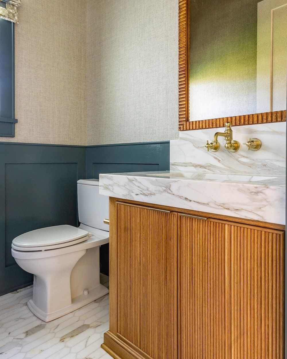 A bathroom with blue paneling, textured wallpaper, a marble vanity with gold hardware, and a white toilet.