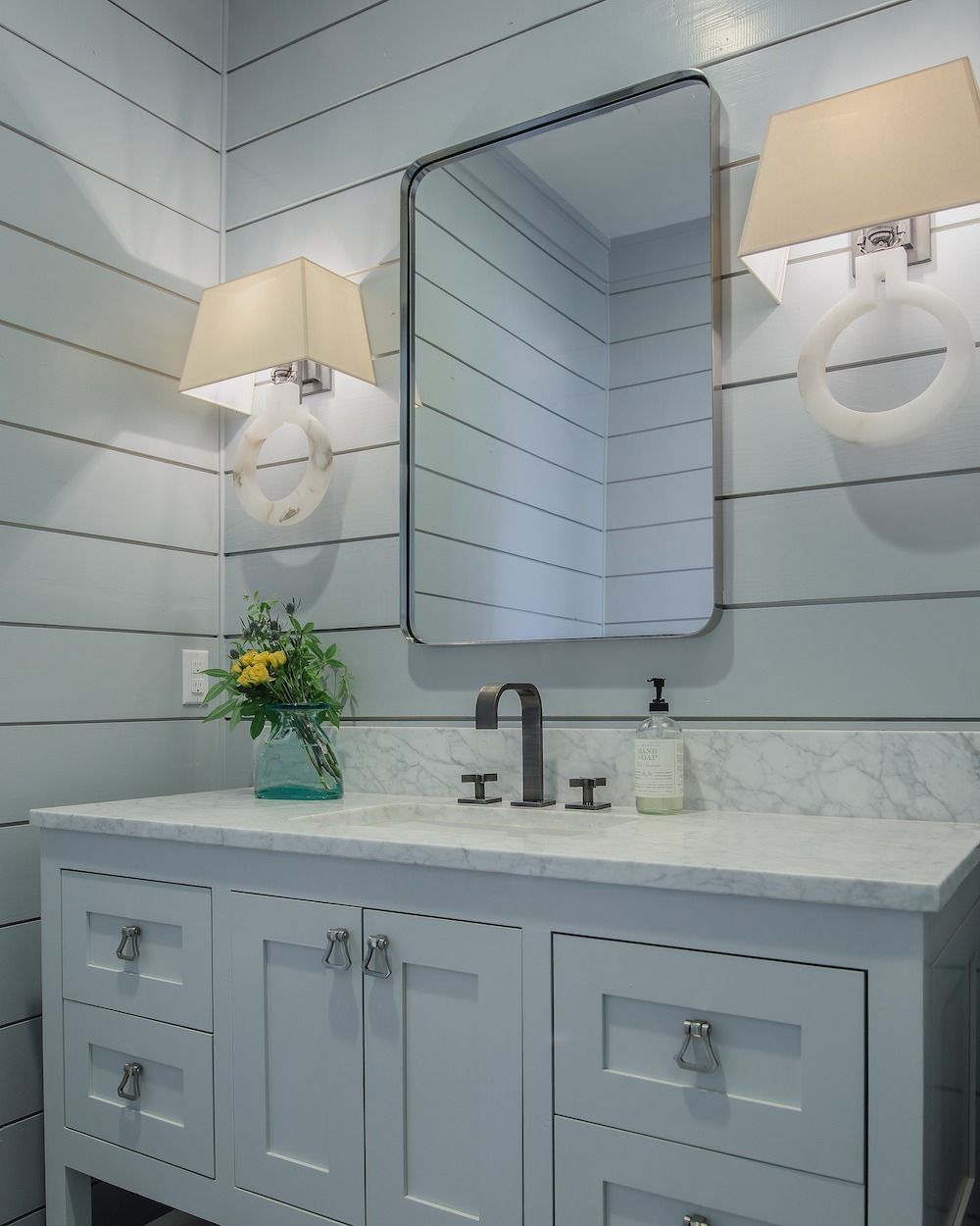 A light blue bathroom with shiplap walls, a vanity, a rectangular mirror, and two beige sconce lamps.