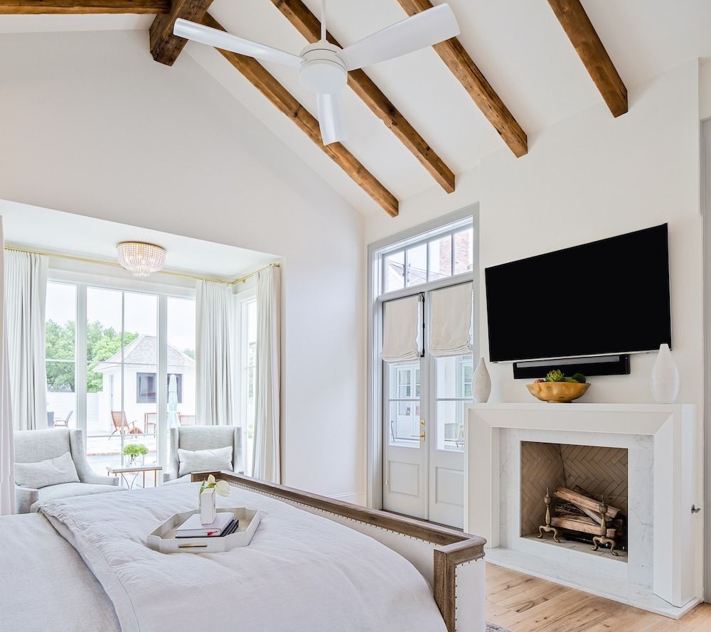 A bright bedroom with a white vaulted ceiling, wooden beams, a ceiling fan, a fireplace with a TV, and a large bed.