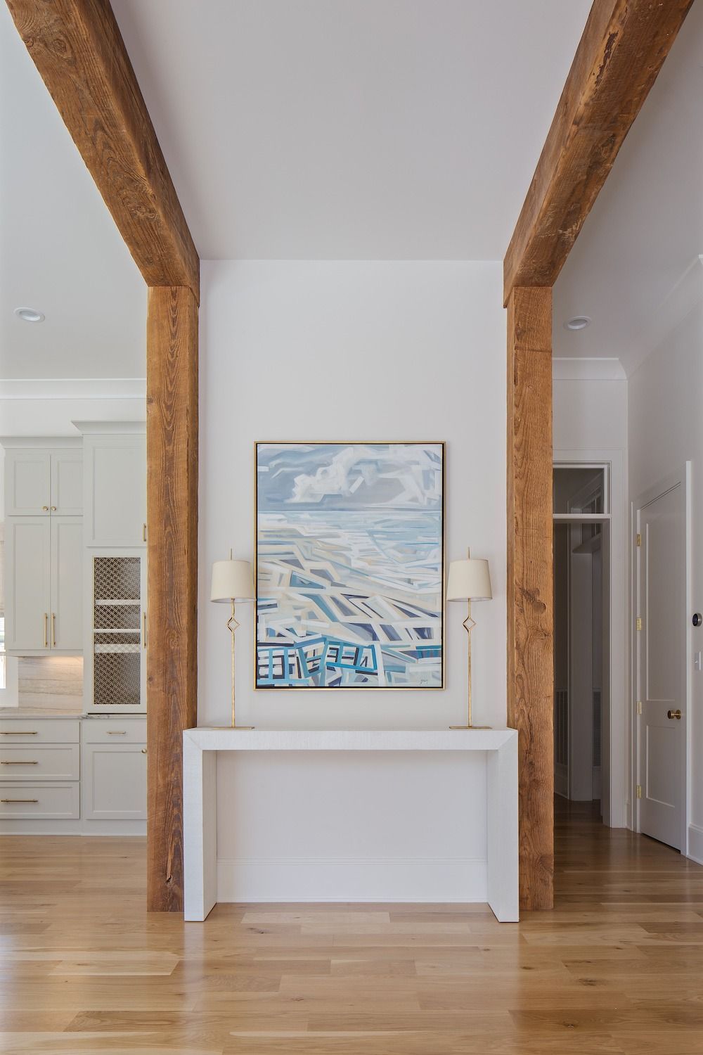 A white console table sits between two vertical wooden beams in a hallway, centered below a large abstract landscape art.