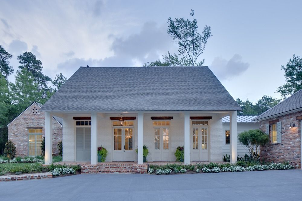 A white cottage with a grey shingled roof, a front porch with white columns, and surrounding greenery at dusk.