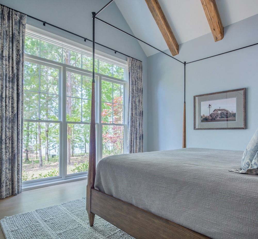 A modern bedroom with a canopy bed, pale blue walls, wood ceiling beams, a large window with patterned curtains, and a rug.