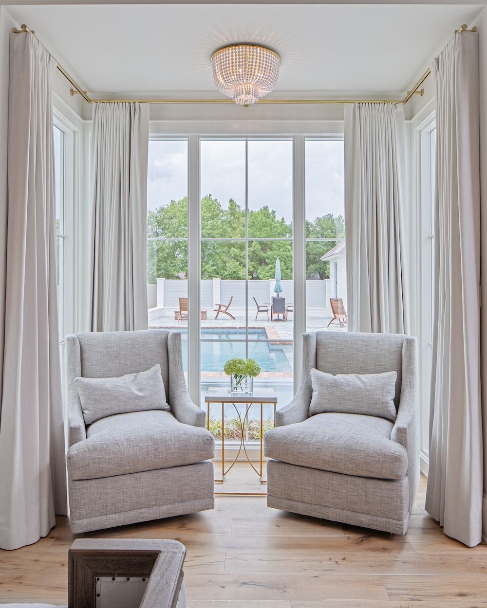 Two light gray armchairs face a large window overlooking a swimming pool, beneath a crystal chandelier.