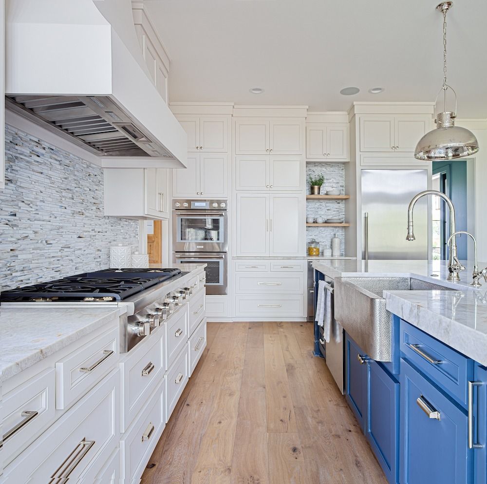 A modern kitchen with white upper cabinets, a marble backsplash, stainless appliances, and blue base cabinets.
