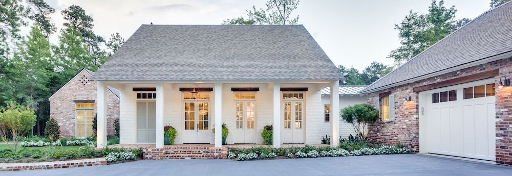 A white, single-story house with a shingled roof, front porch, and attached brick garage in a wooded setting.