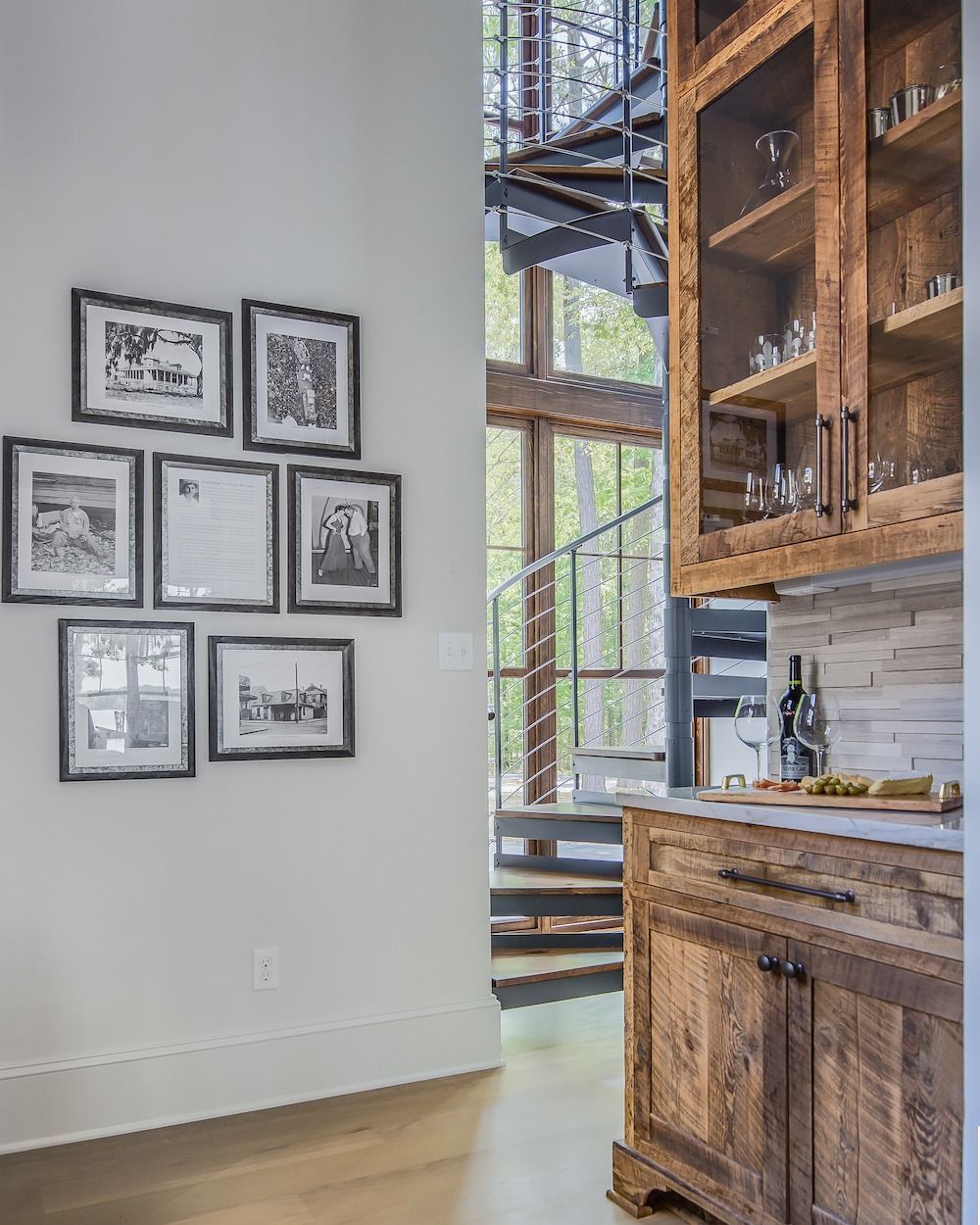 A wood bar cabinet with a glass hutch sits next to a gallery wall of seven black-and-white framed photos near a spiral stair.