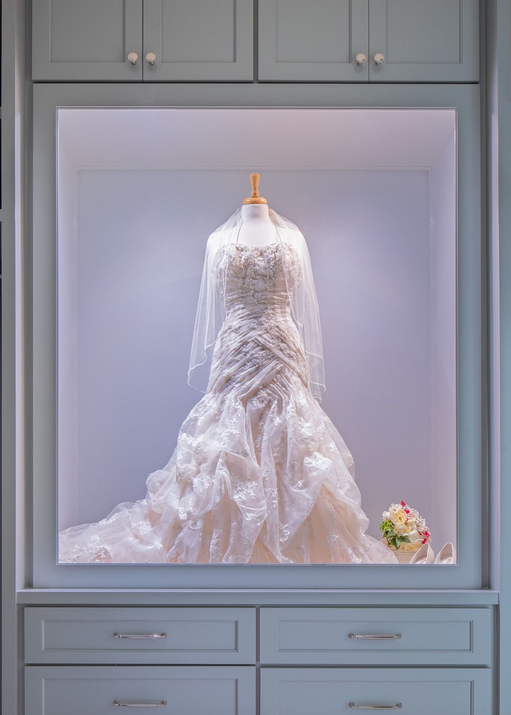 A white, ornate wedding gown displayed on a mannequin inside a built-in illuminated cabinet with a small floral bouquet.