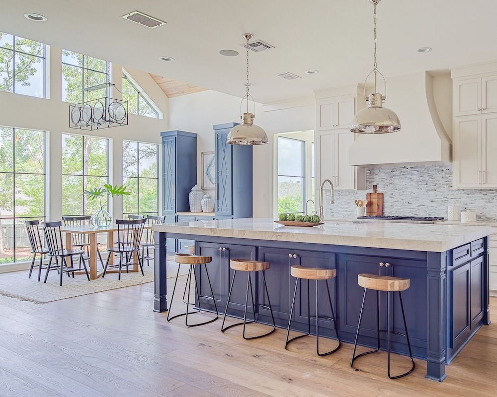 A modern kitchen with a large navy blue island, wooden bar stools, white cabinets, and a dining area with floor-to-ceiling windows.