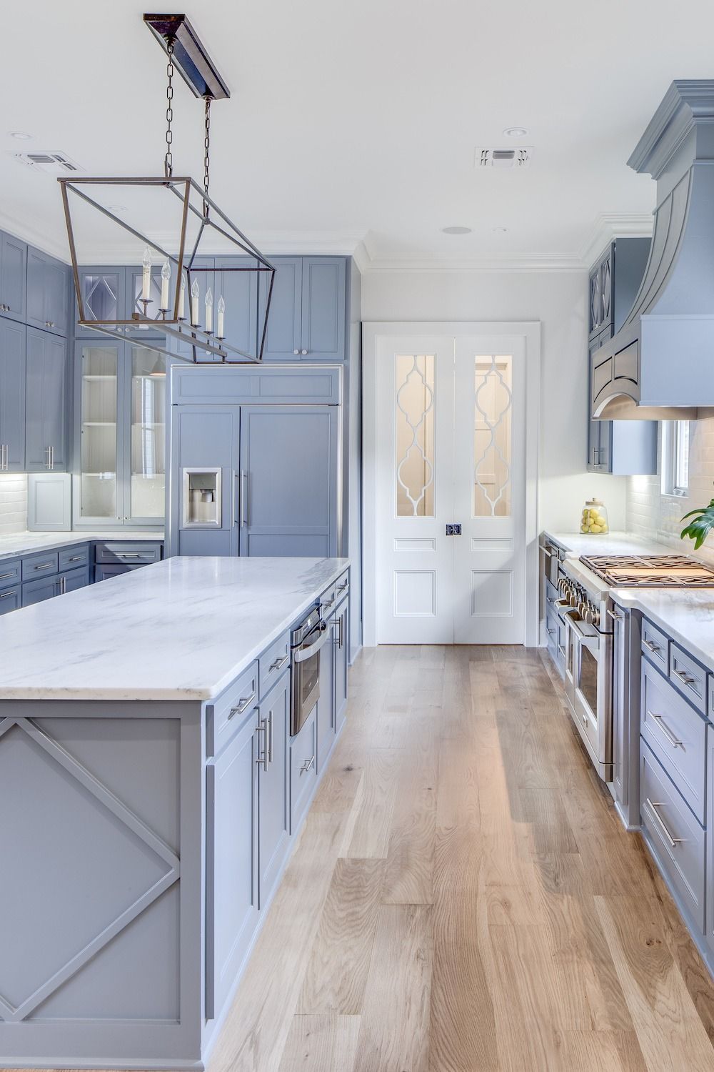 A modern kitchen with light blue cabinets, a white marble island, stainless steel appliances, and wood flooring.