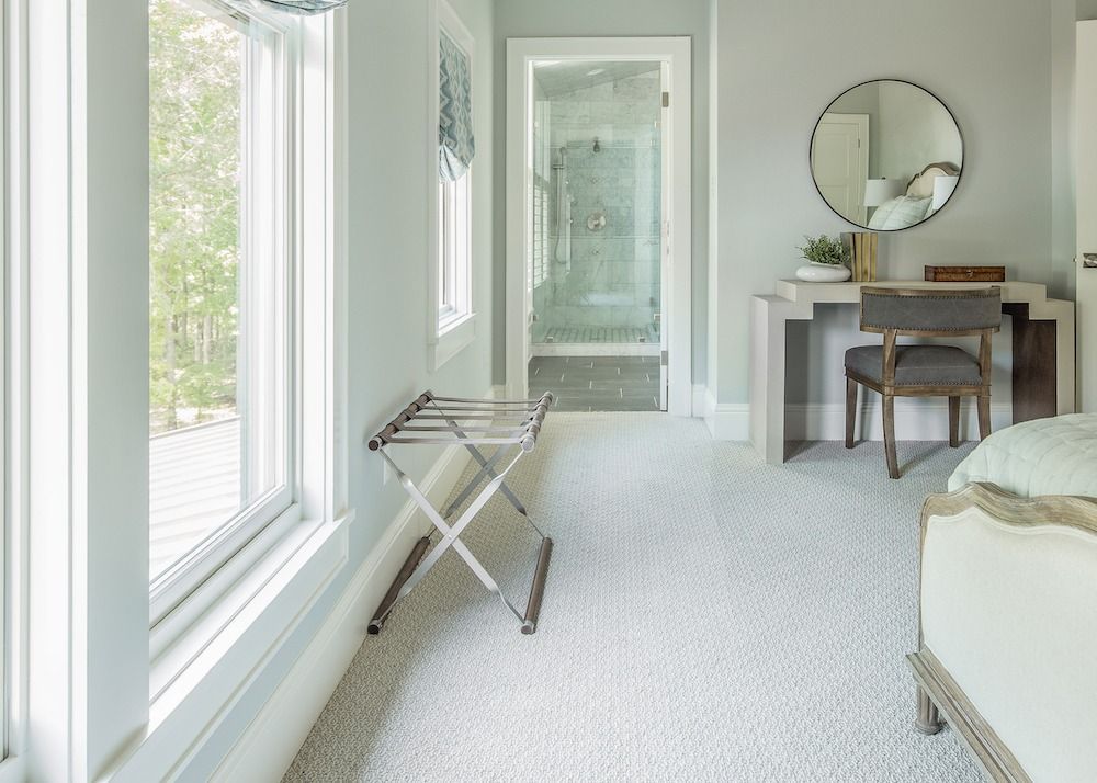 A light-colored bedroom features a metal luggage rack, a vanity with a circular mirror, and a view into a bathroom.