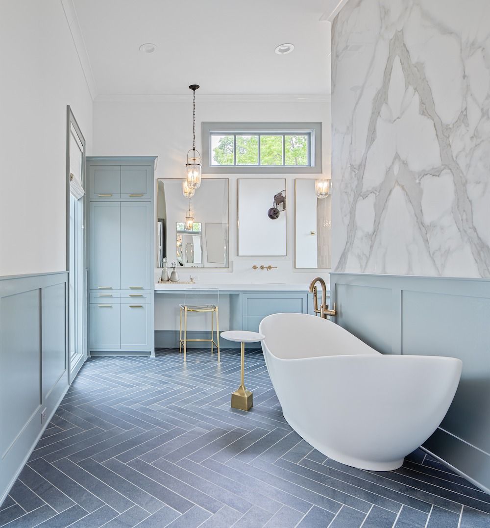 A spacious, serene bathroom features a white soaking tub, blue cabinetry, herringbone tile floors, and marble accents.