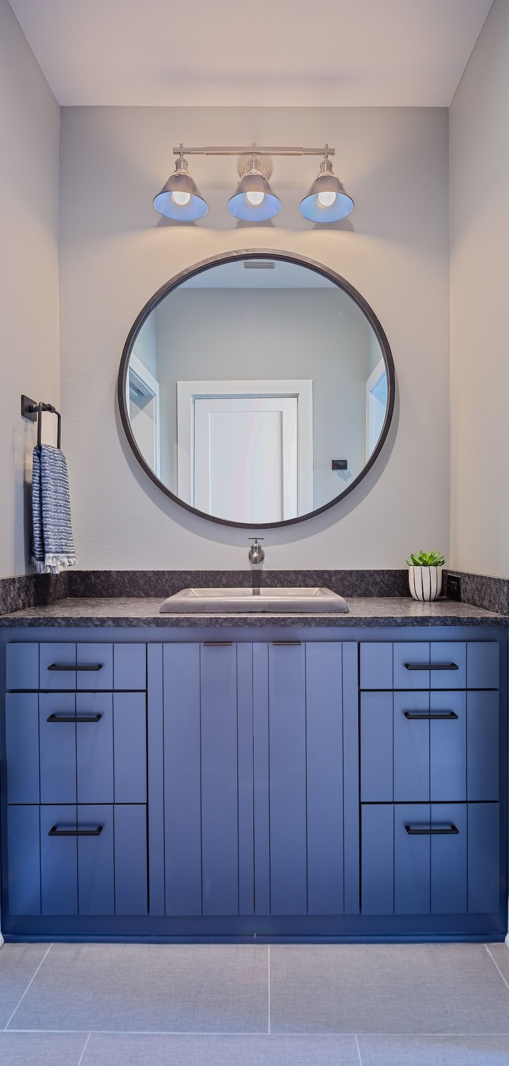 A modern bathroom vanity with navy blue cabinetry, a round mirror, and a three-bulb light fixture.
