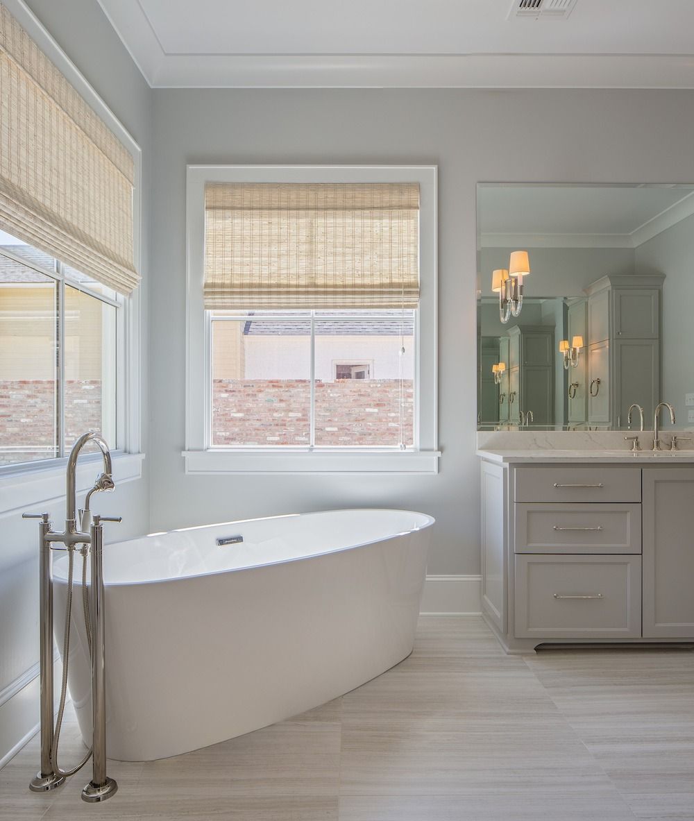 A modern bathroom featuring a white freestanding tub, a grey vanity, and woven window shades under soft lighting.