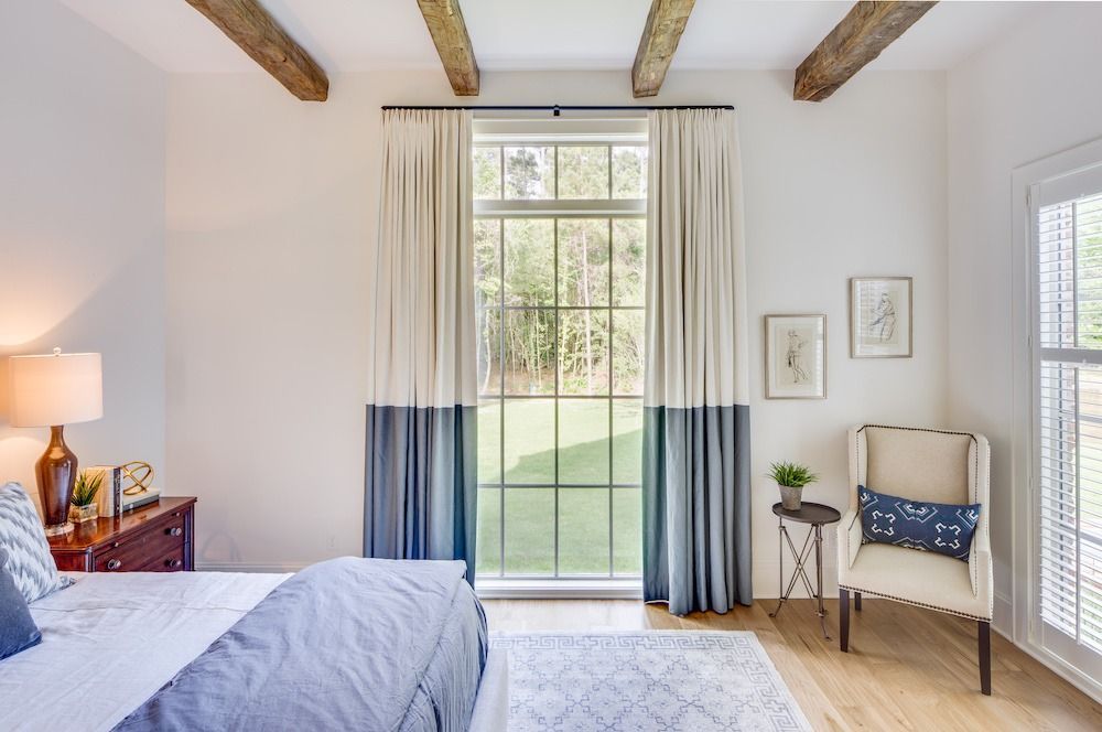 A bright bedroom featuring a tall window with two-toned curtains, a bed, and an armchair near a side table.