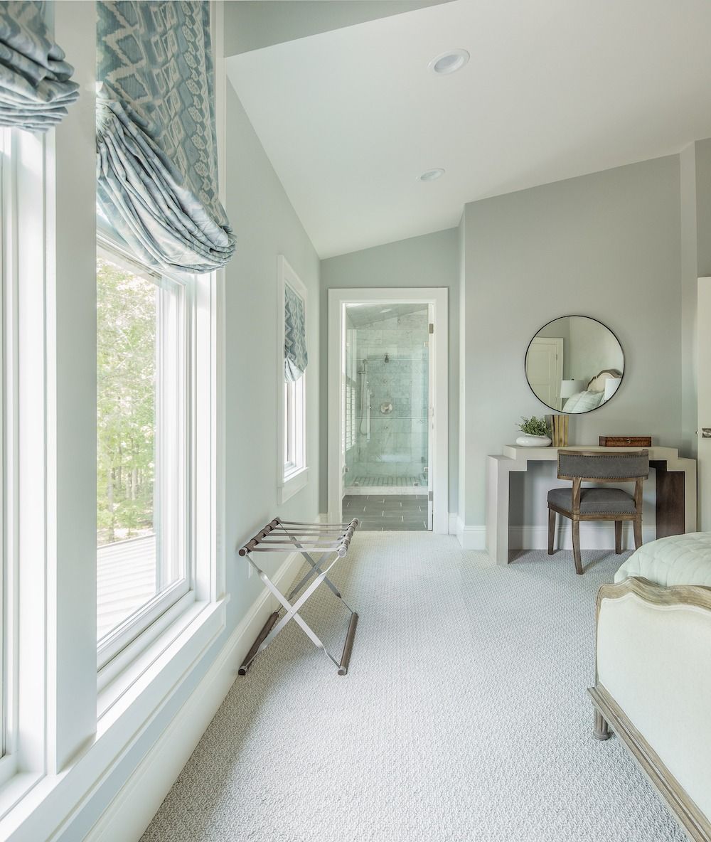A bright, neutral-toned bedroom features a small desk, round mirror, and a suitcase rack by a window with blue shades.