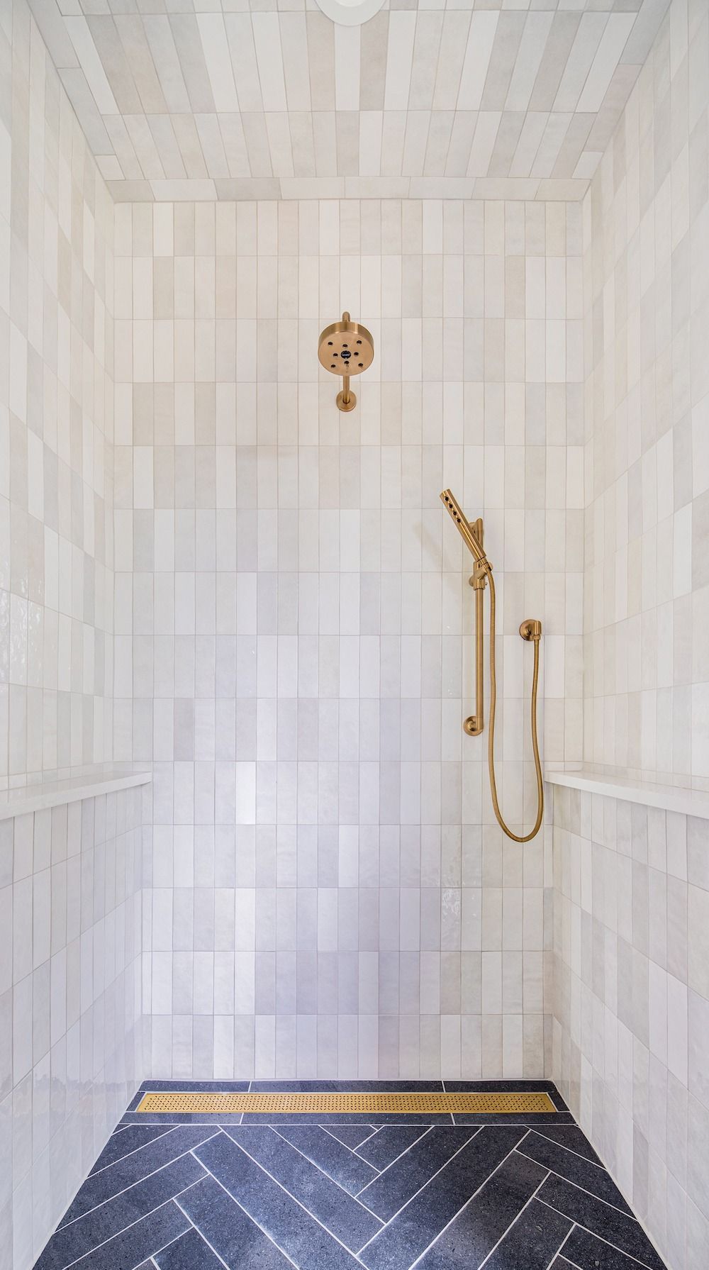 A modern shower with white vertical tiles, brass fixtures, a handheld sprayer, and a dark herringbone-patterned floor.