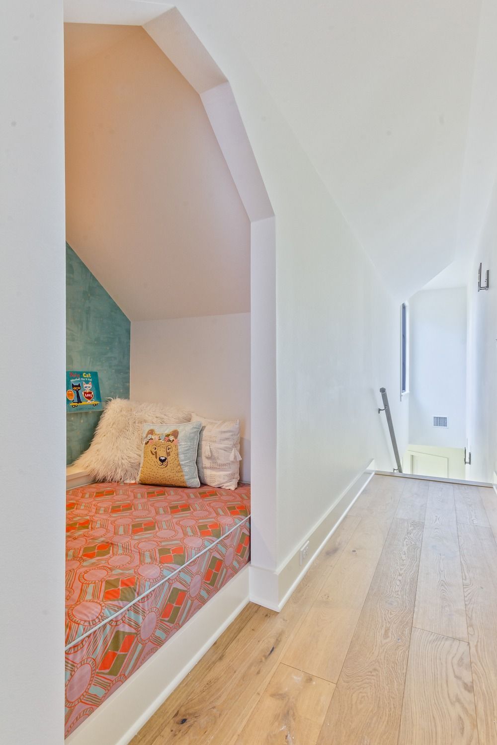 A cozy, arched reading nook with patterned bedding and a plush fox pillow, set off a bright hallway with hardwood floors.