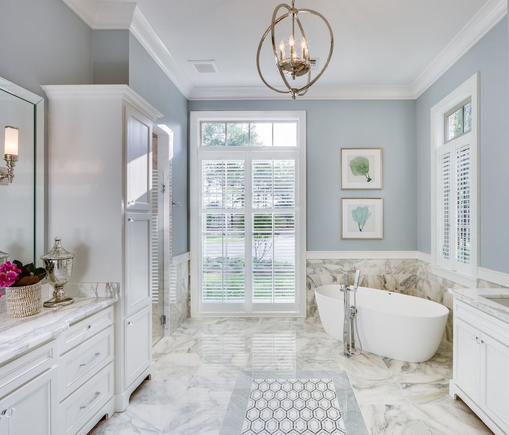A bright bathroom with white cabinetry, marble flooring, a freestanding oval tub, and light blue walls.