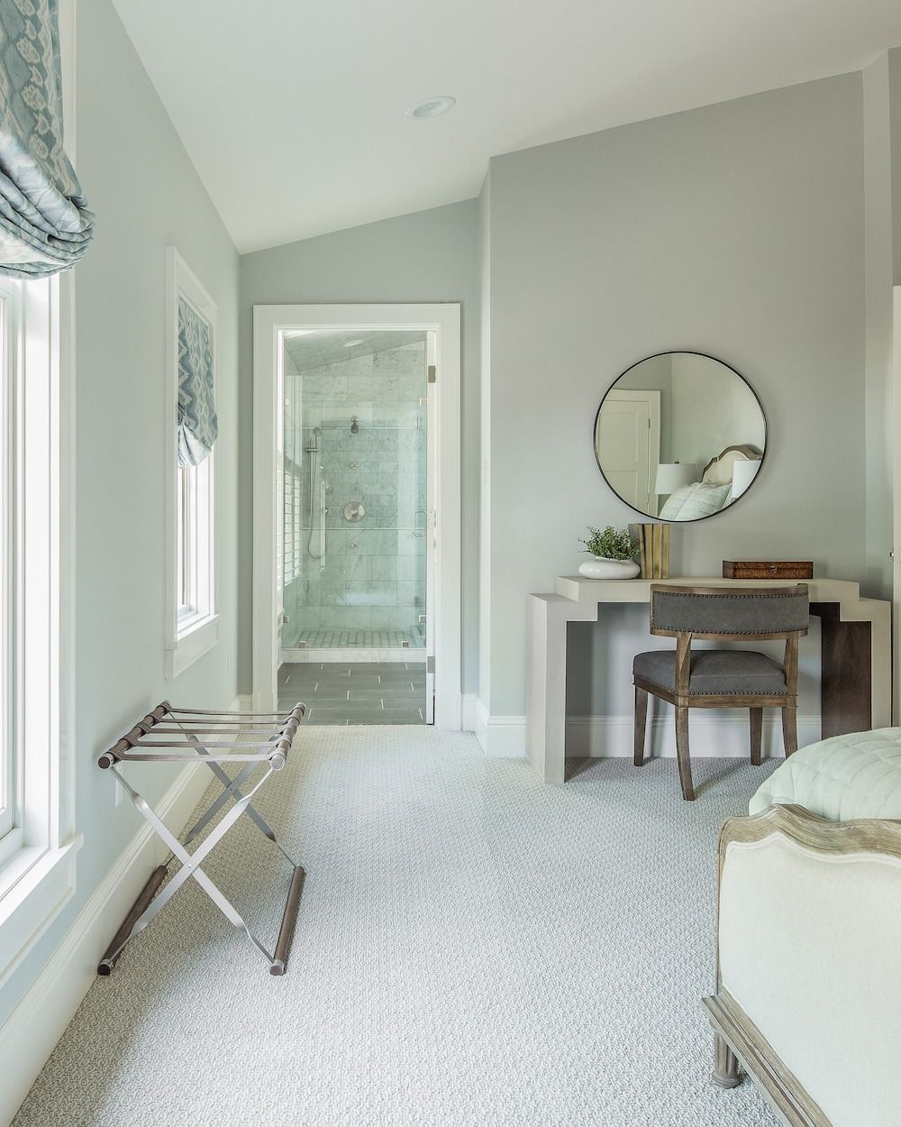 A light-colored bedroom with a luggage rack, a desk with a round mirror, and a doorway leading to a tiled bathroom.