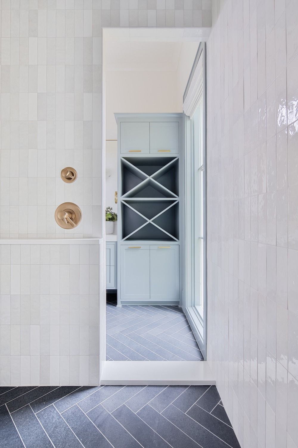 A bathroom with light-gray tile walls, brass shower fixtures, and a herringbone floor looking into a room with wine racks.